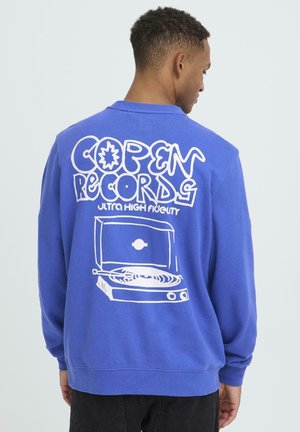 Junger Mann trägt einen blauen Sweatshirt mit weißer Grafik und dem Text "Copen Records Ultra High Fidelity" sowie einer Zeichnung eines Plattenspielers auf der Rückseite.