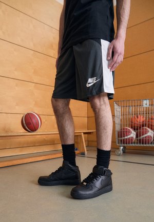 Osoba ubrana w czarne spodenki Nike i czarne buty sportowe Nike stoi na podłodze siłowni obok piłek do koszykówki i drewnianej ławki.
