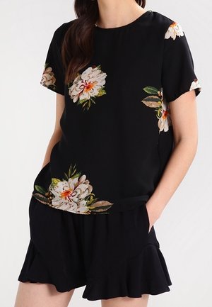 Sort kortærmet top med blomsterprint i hvid, gul og grøn. Glat stof, rund halsudskæring og afslappet pasform. Parret med sorte shorts med volanger.