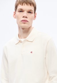 Kremsa krāsas kokvilnas polo krekls ar apkakles izgriezumu, divu pogu rāvējslēdzēju un mazu sarkanu "cK" logotipu kreisajā krūtīs.