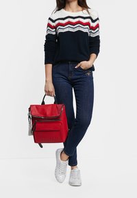 Roter Leder-Rucksack mit einem Reißverschluss oben, schwarzen Akzenten und einem Quast-Detail. Das Modell trägt einen marineblauen Pullover mit roten und weißen Zickzackstreifen, blaue Jeans und silberne Sneaker.