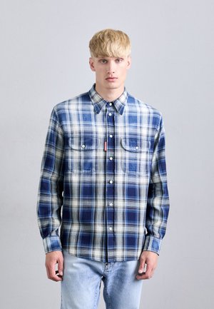 Ung mand med kort blond hår iført en blå og hvid rutet skjorte med knapper og lyseblå jeans, stående mod en ensfarvet grå baggrund.