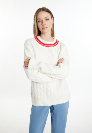 Giovane donna con capelli lisci castano chiaro indossa un maglione bianco a trecce con strisce rosse e jeans azzurri, braccia incrociate.