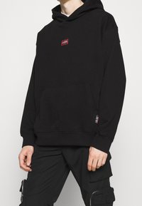 Sweat-shirt à capuche noir avec une poche avant, arborant un écusson logo carré rouge sur la poitrine. Fabriqué en tissu doux et texturé.