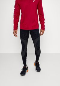 Chemise rouge à manches longues et leggings noirs avec des motifs texturés, dotés d'accents réfléchissants. Chaussures de sport noires avec des détails orange.
