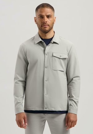 Hombre de frente vistiendo una chaqueta abotonada de color gris claro con un bolsillo en el pecho y pantalones a juego contra un fondo liso.