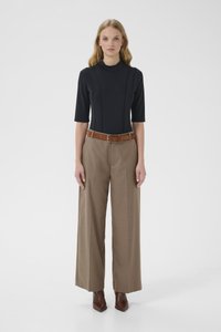 Noor naine seisab mustas istuvas toppis, laia säärega beežides pükstes pruuni vööga ja pruunides teravnurksetes saapades valge tausta ees.