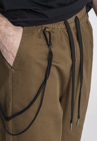 Bruine cargo-broek met een tailleband met trekkoord. Kenmerken zijn zwarte trekkoorden en metalen uiteinden, met zijzakken en een gladde stofstructuur.