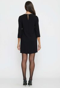 Schwarzes, knielanges Kleid mit dreiviertel Ärmeln, Schlüsselrücken-Detail und einem mit Perlen verzierten Ausschnitt. Durchsichtige schwarze Strumpfhosen und schwarze High Heels.