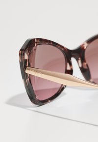 Óculos de sol de plástico com uma armação marrone marmorizada e lentes rosa degradê. Apresenta um formato angular de olhos de gato e hastes de metal dourado com o logotipo.