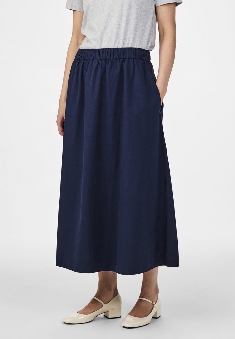 Tmavě modrá midi sukně s elastickým pasem, s bočními kapsami a hladkou texturovanou látkou. Doplněná krémově zbarvenými botami.