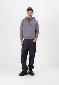 Sudadera gris con un bolsillo frontal tipo canguro y cordones, combinada con jeans baggy oscuros y amplios que cuentan con una etiqueta verde visible. Zapatos negros gruesos.