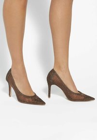Brown stiletto heels made of mesh material, adorned with small metallic studs, featuring a pointed toe and slim high heel for an elegant silhouette.