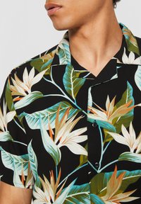 Bărbat purtând o cămașă neagră cu mâneci scurte, cu imprimeu colorat de floare tropicală pasăre paradis și frunză verde, guler deschis, cu fața spre stânga.