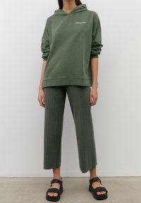 Sudadera con capucha de color verde oliva con un logo frontal, combinada con pantalones cortos y acanalados en verde oliva. Sandalias negras gruesas completan el conjunto.