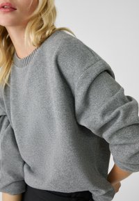 Grauer Strickpullover mit Rundhalsausschnitt, überschnittenen Schultern und langen Ärmeln; weiche Textur und dezente Rippendetails am Ausschnitt.