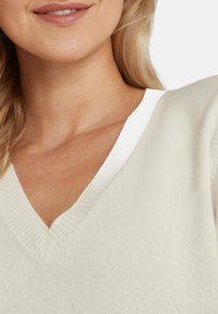 Maglione a V in crema chiaro, con texture a coste, collo bianco a contrasto e tessuto morbido. Primo piano del dettaglio del collo.