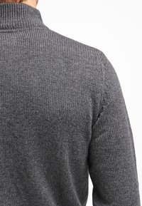 Pull en tricot gris avec un col montant. Le tissu présente un motif texturé, mettant en valeur son apparence douce et chaude.