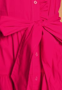 Ljusrosa klänning med framknappar och en stor knut i midjan, med synliga sömmar och volangdetaljer.