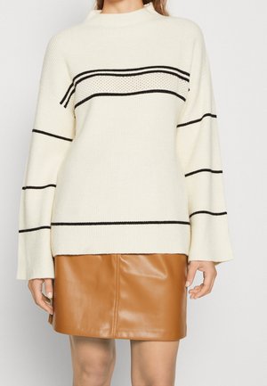 Woman wearing a cream sweater with black horizontal stripes and a brown leather skirt standing against a plain background.