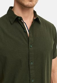 Kurzärmliges, olivgrünes Button-Down-Hemd aus strukturiertem Stoff; mit einem gemusterten Innenkragen und gestreiften Details; fünf sichtbare Knöpfe.