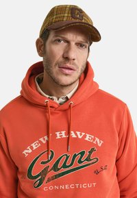 Oranje hoodie met groene grafische logo "NEW HAVEN Gant CONNECTICUT" met trekkoordjes. Bruine geruite pet met de letter "G" aan de voorkant.