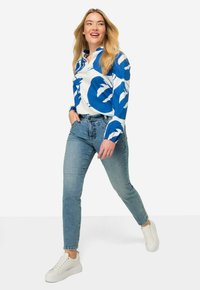 Långärmad blus med stort blått och vitt blommönster; kombinerad med ljusblå jeans och vita sneakers. Slät tygstruktur.