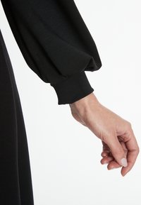 Schwarzes Strickkleid mit voluminösen Ärmeln und eng anliegenden Manschetten, das eine glatte Textur und eine schlanke Form zeigt. Hand in der Nähe des Ärmels positioniert.