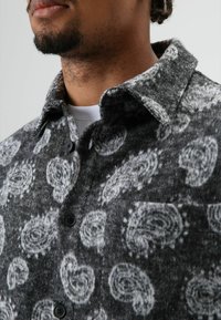 Fleece shirt in grijs met zwarte spiraalpatronen, voorzien van een knoopsluiting aan de voorkant en een kraag. Zachte textuur en licht gestructureerde stof.