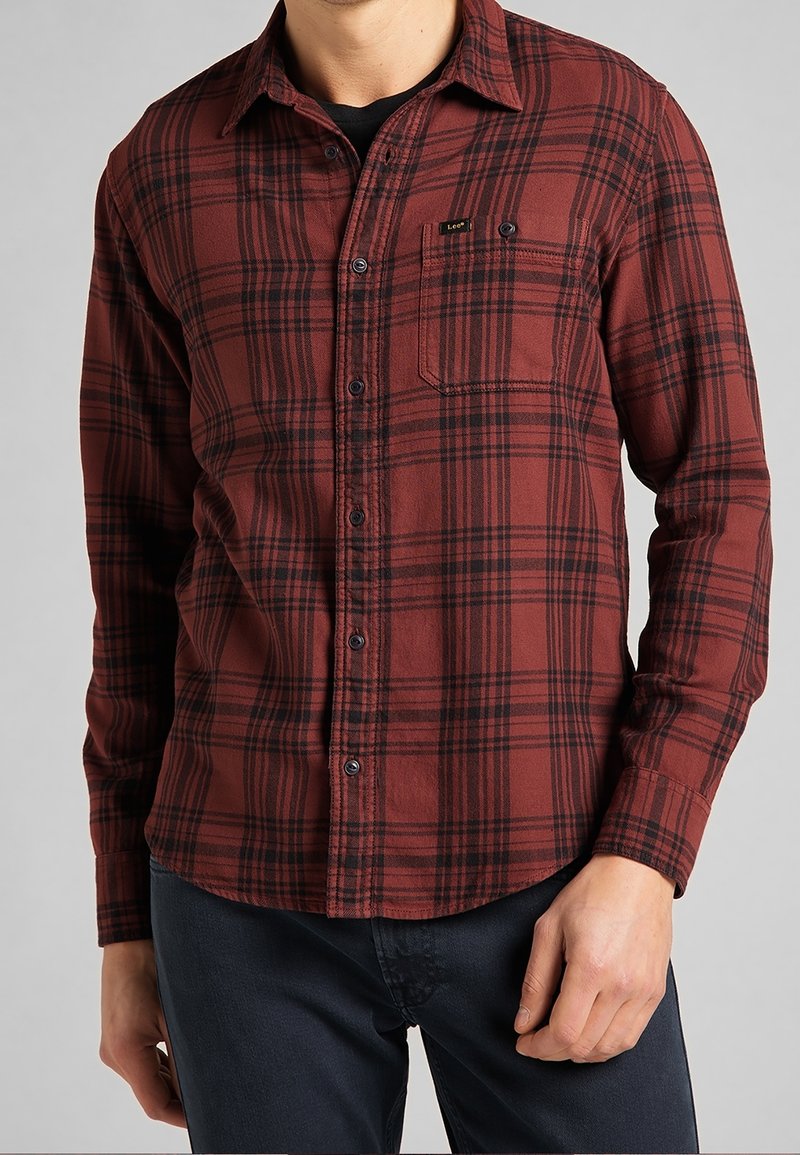Camisa de franela a cuadros rojos y negros con mangas largas, cierre de botones y bolsillo en el pecho. Textura suave, corte relajado y bajo curvado.