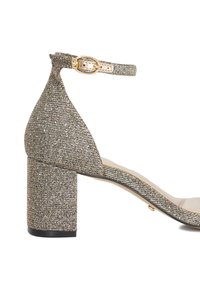 Glittery silver block-heeled sandal with a pointed toe and ankle strap featuring a gold buckle. Textured fabric adds visual interest.