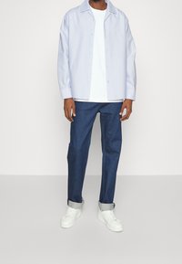 Camicia azzurra chiara con bottoni, T-shirt bianca, pantaloni in denim scuri e sneaker bianche. Abbigliamento casual con una vestibilità comoda e maniche arrotolate.