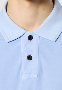 Polo bleu clair en tissu texturé, doté d'un col classique et de deux boutons noirs à l'encolure.