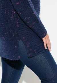 Granatowy sweter dzianinowy z różowymi plamkami, z prążkowaną teksturą i bocznymi rozcięciami. W zestawie z dopasowanymi ciemnoniebieskimi dżinsami.