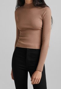 Femme portant un haut ajusté à manches longues côtelé de couleur camel et un pantalon taille haute noir, posant devant un fond gris uni.