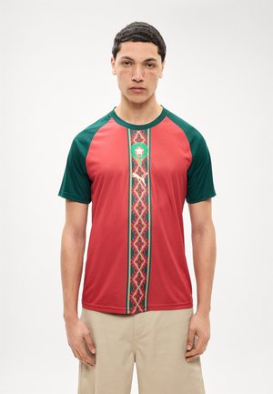 Uomo che indossa una maglia da calcio a maniche corte rossa e verde con una striscia verticale a motivo e pantaloni beige, in piedi su uno sfondo bianco.