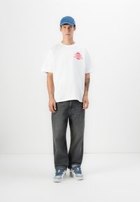 Hvit bomulls T-skjorte med rødt grafikk, grå baggy jeans, og blå denim caps. Sneakers er lyseblå med mørkeblå snøringer. Enkel bakgrunn.