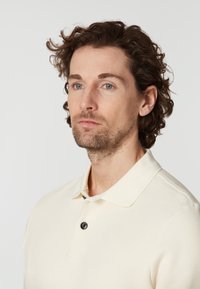 Manskortärmad polo i off-white, med en strukturerad tygkvalitet, klassisk krage och en enda svart knapp vid halsen.