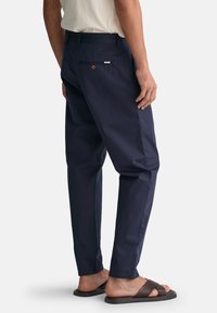 Marineblå bomuldsbukser med slim fit og bæltestropper, der har en baglomme med knap og læderdetalje. Sammen med brune sandaler.