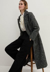Cappotto bianco e nero testurizzato con design doppiopetto, abbinato a una blusa bianca e pantaloni neri a gamba larga, seduto su uno sgabello di legno.