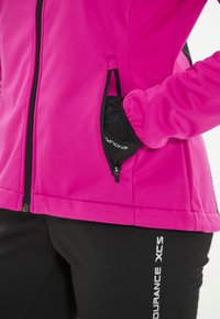 Fuchsia softshell-jakke med sorte accenter, med en lynlås-lomme. Båret med sorte tætsiddende bukser, der viser side-logoet.