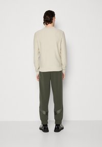 Suéter de punto crema combinado con pantalones oscuros verdes ajustados que presentan detalles brillantes, vistos desde la parte de atrás contra un fondo liso.