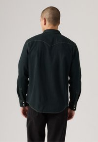 Dark green denim shirt with a western-style yoke, buttoned cuffs, and a curved hem, displayed from the back. Textured fabric, casual fit.