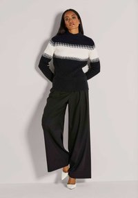 Frau, die einen schwarz-weiß gestreiften Rippstrickpullover, weit geschnittene schwarze Hose und weiße spitze Pumps trägt und an einer schlichten Wand steht.