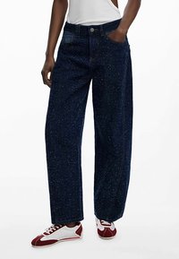 Tmavě modré džíny s širokými nohavicemi a tečkovanou texturou, s předními kapsami a zapínáním na knoflíky, doplněné bílými a červenými teniskami.