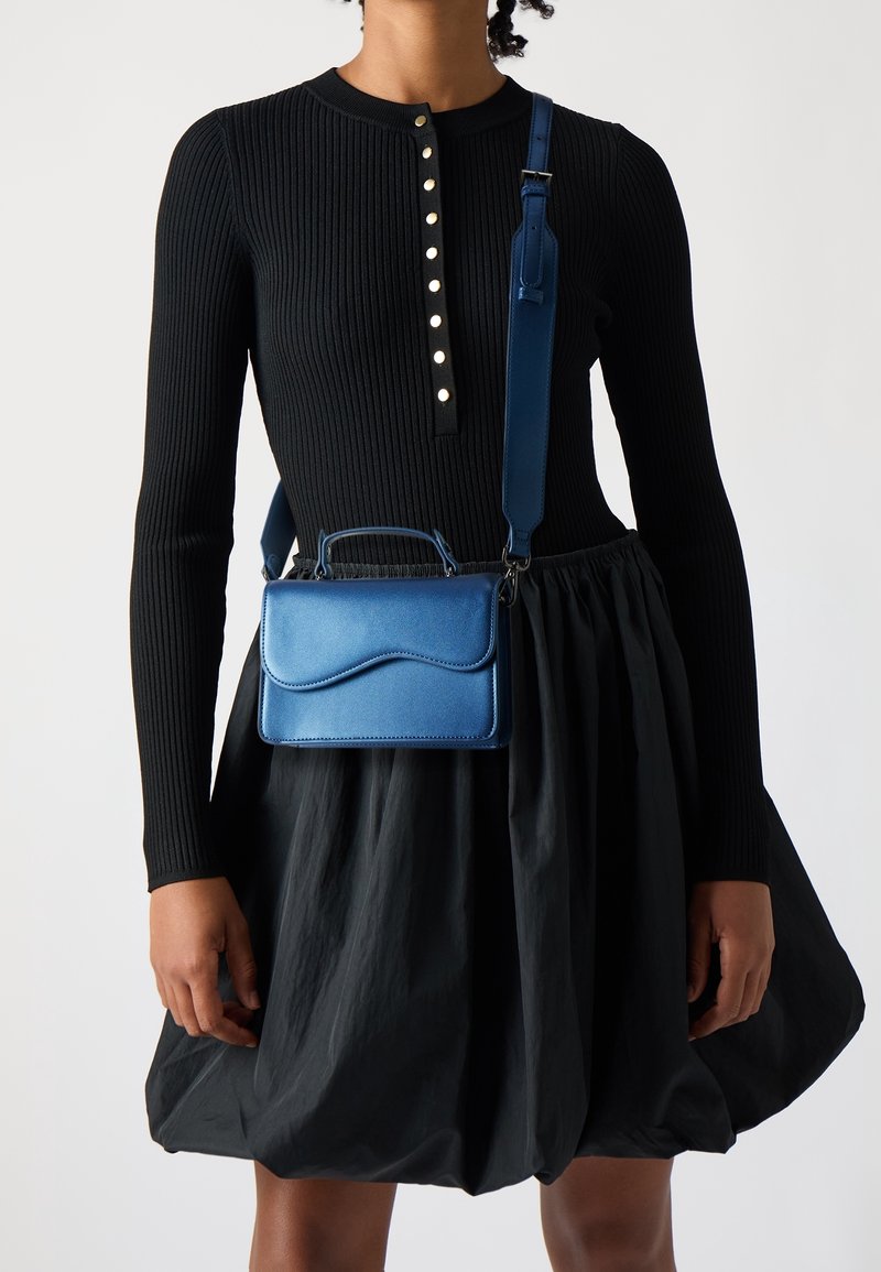 Blå taske i kunstlæder med buet klap, topgreb og justerbar skulderrem, vist mod en sort ribbet top og en flydende sort nederdel.