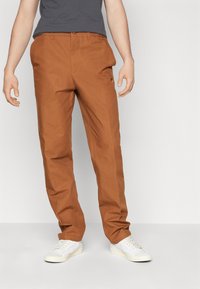 Pantalones marrones de corte recto, fabricados con una mezcla de algodón, con bolsillos inclinados y un sutil logo en el lado, combinados con zapatillas blancas.