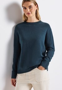 Pullover aus dunkelblauem Strickstoff mit Rundhalsausschnitt, gerippten Bündchen und Saum; getragen mit hellbeigen Hosen, die eine entspannte Passform und Textur zeigen.