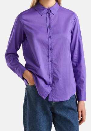 Osoba ubrana w fioletową, zapinaną na guziki koszulę z długim rękawem i kołnierzykiem, z ręką w kieszeni, w połączeniu z niebieskimi dżinsami.