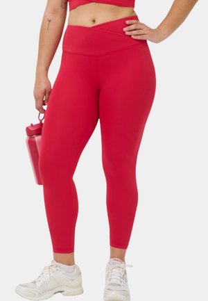 Vrouw draagt rode leggings met hoge taille en witte sneakers, houdt in één hand een bijpassende rode waterfles vast, staat met een hand op haar heup.